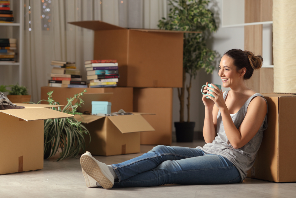 A woman sitting on the floor around boxes, drinking coffee